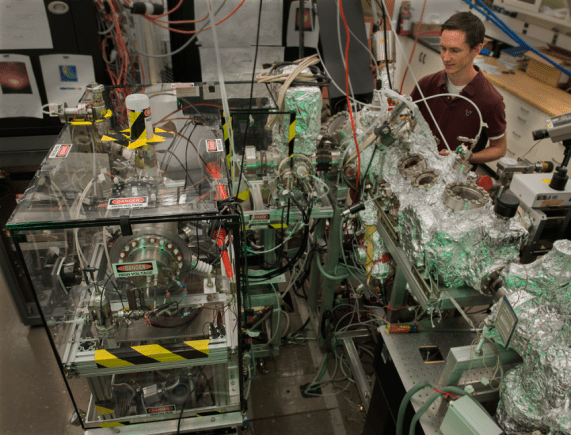 NIST researcher Kevin Dwyer with the silicon enrichment apparatus. The process begins at the left, where natural-abundance silicon in the form of silane gas (SiH4) is ionized. The ions pass through a magnetic field (top left), which causes their paths to curve to different degrees based on their mass. The silicon-28 ions, now separated from the other silicon isotopes, are decelerated through an ultra-high vacuum chamber (top center) and into the deposition chamber (top right) (Credit: Pomeroy, NIST, Dwyer)